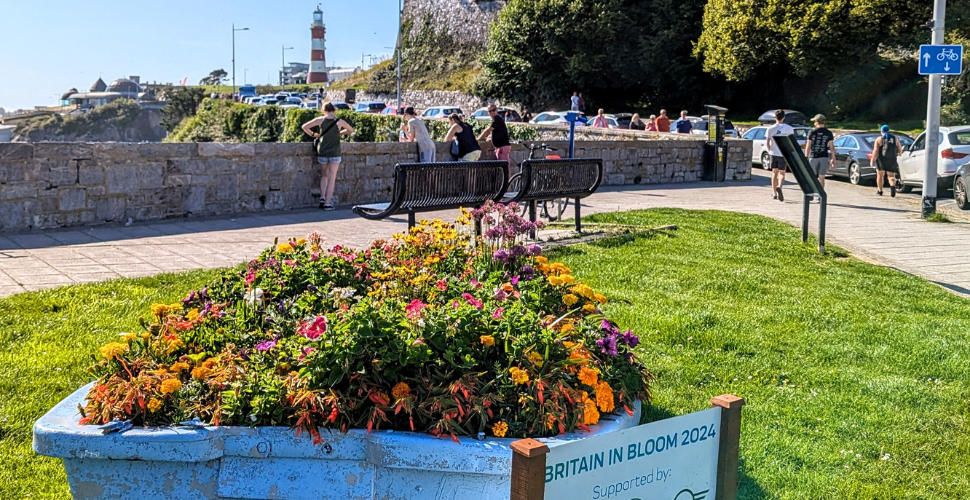 A boat full of flowers with cars and a road on the right with Smeaton's Tower in the background